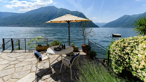 Un patio con mesa, sillas y sombrilla con vista a un lago y montañas al fondo.