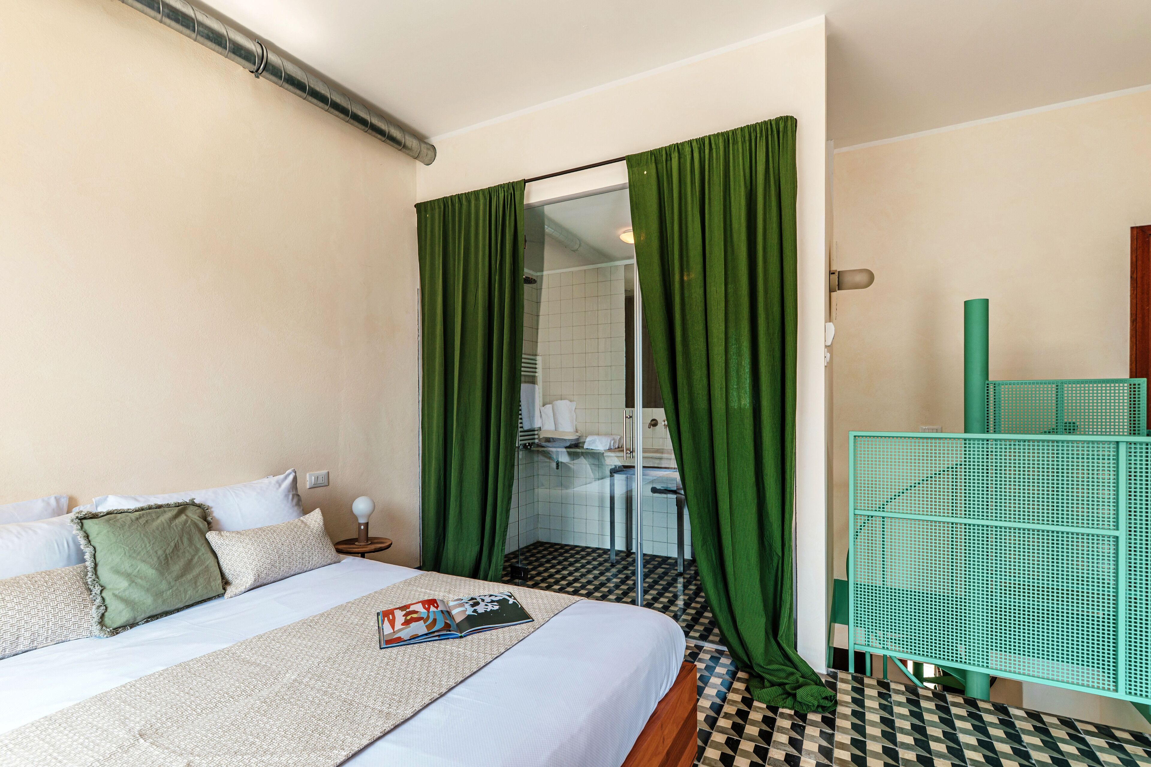 Een hotelkamer met een bed, groene gordijnen, een geruit vloerkleed en een badkamer zichtbaar door een glazen deur.