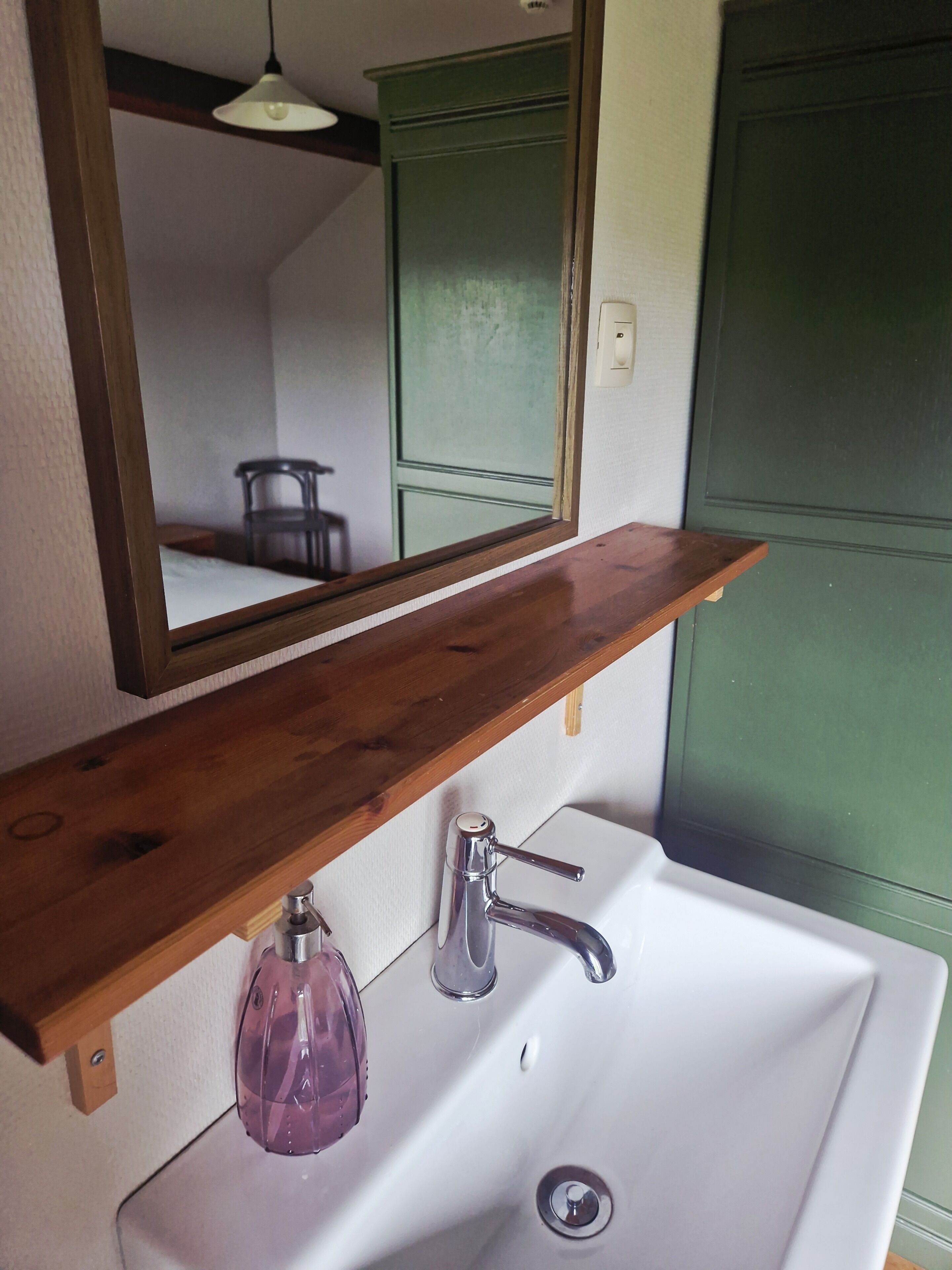 A bathroom with a wooden shelf above a white sink, a mirror, and a green cabinet.