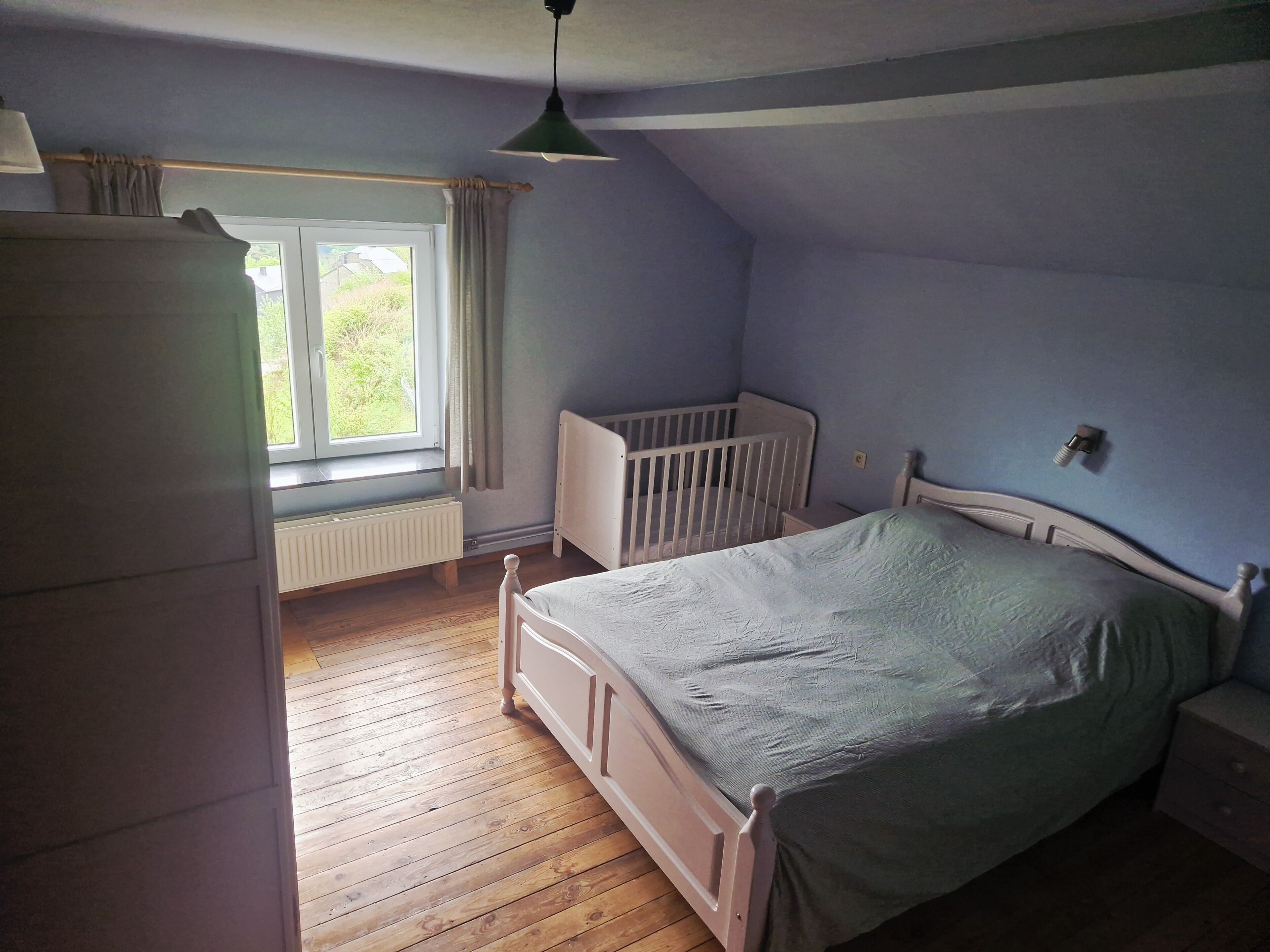 Une chambre avec un plancher en bois, un berceau blanc, un lit recouvert d’une couverture verte, une fenêtre avec des rideaux et un radiateur.