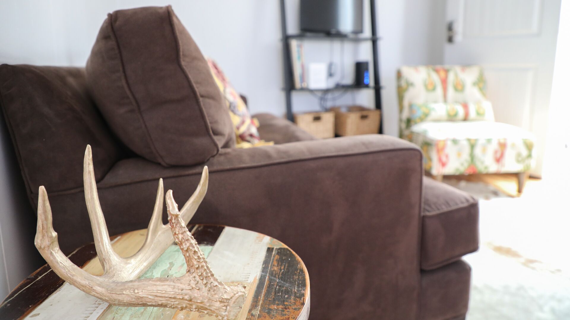 Un salon avec un canapé marron, un objet décoratif en forme de bois de cerf posé sur une table, et une chaise colorée à l’arrière-plan.