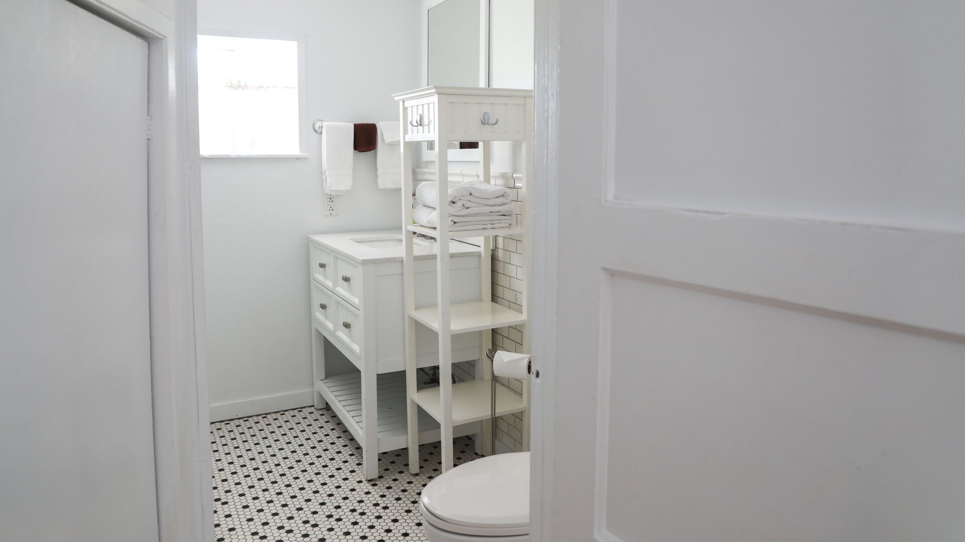 Um banheiro pequeno e branco, com vaso sanitário, toucador branco com gavetas, prateleira com toalhas e espelho.