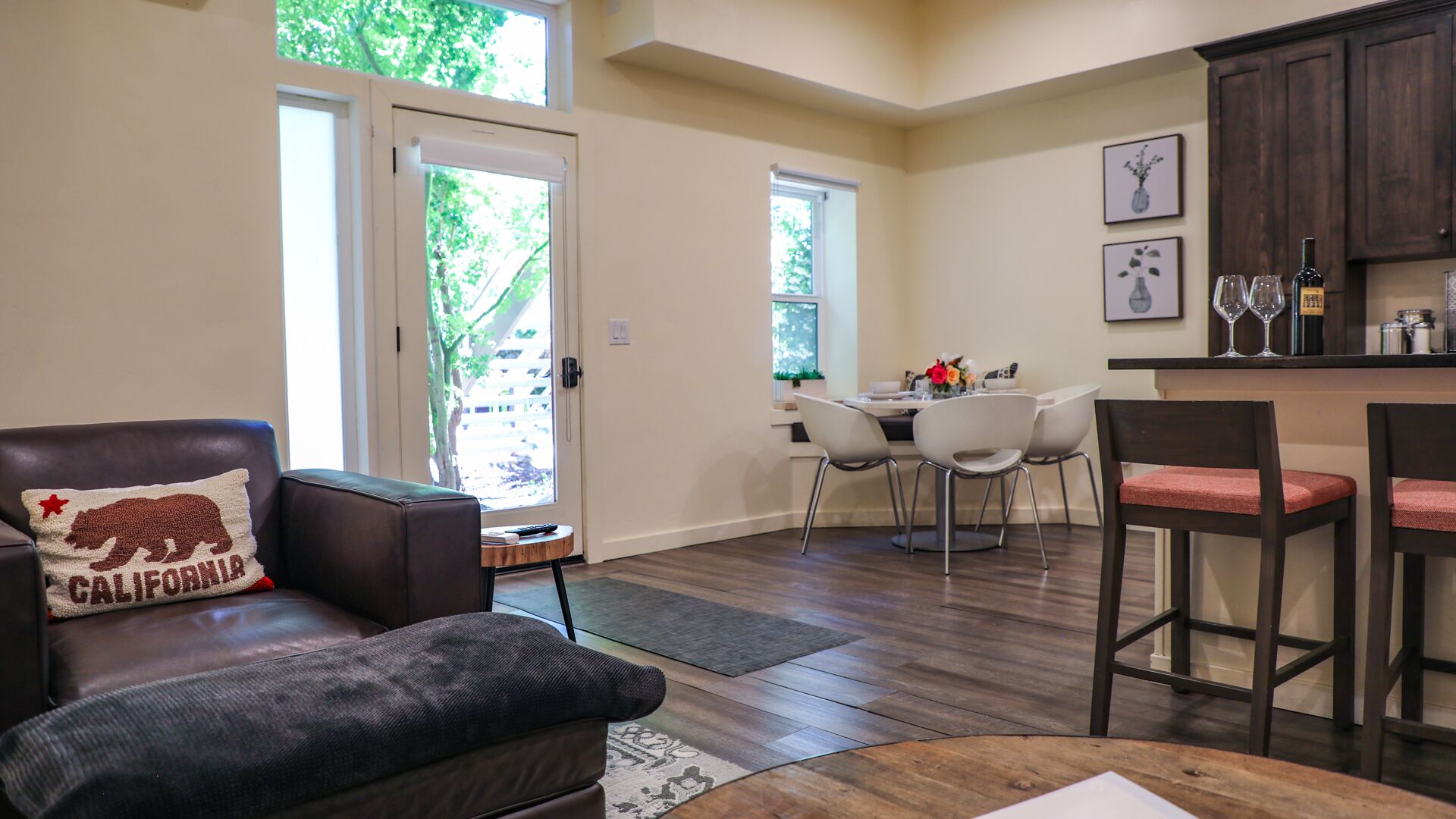 A living room with a leather chair, a dining area with a table and chairs, a kitchen area with bar stools, and a sliding glass door leading to a patio.