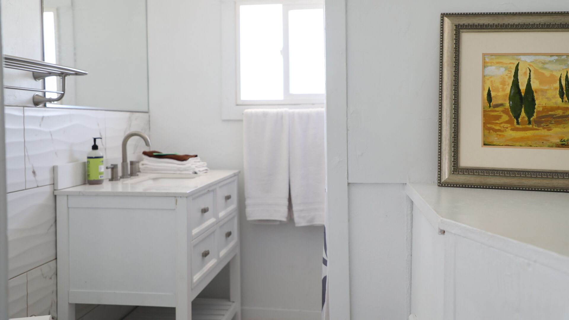 Un baño con tocador blanco, un espejo, una ventana y un cuadro enmarcado en la pared.