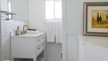 Un baño con tocador blanco, un espejo, una ventana y un cuadro enmarcado en la pared.