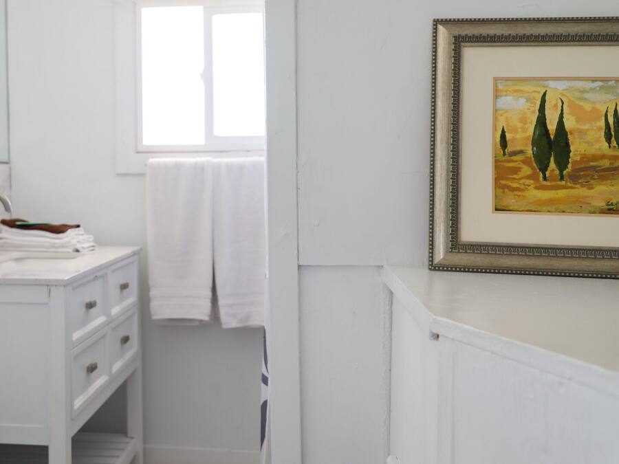 Une salle de bain avec un meuble-lavabo blanc, un miroir, une fenêtre et un tableau encadré au mur.