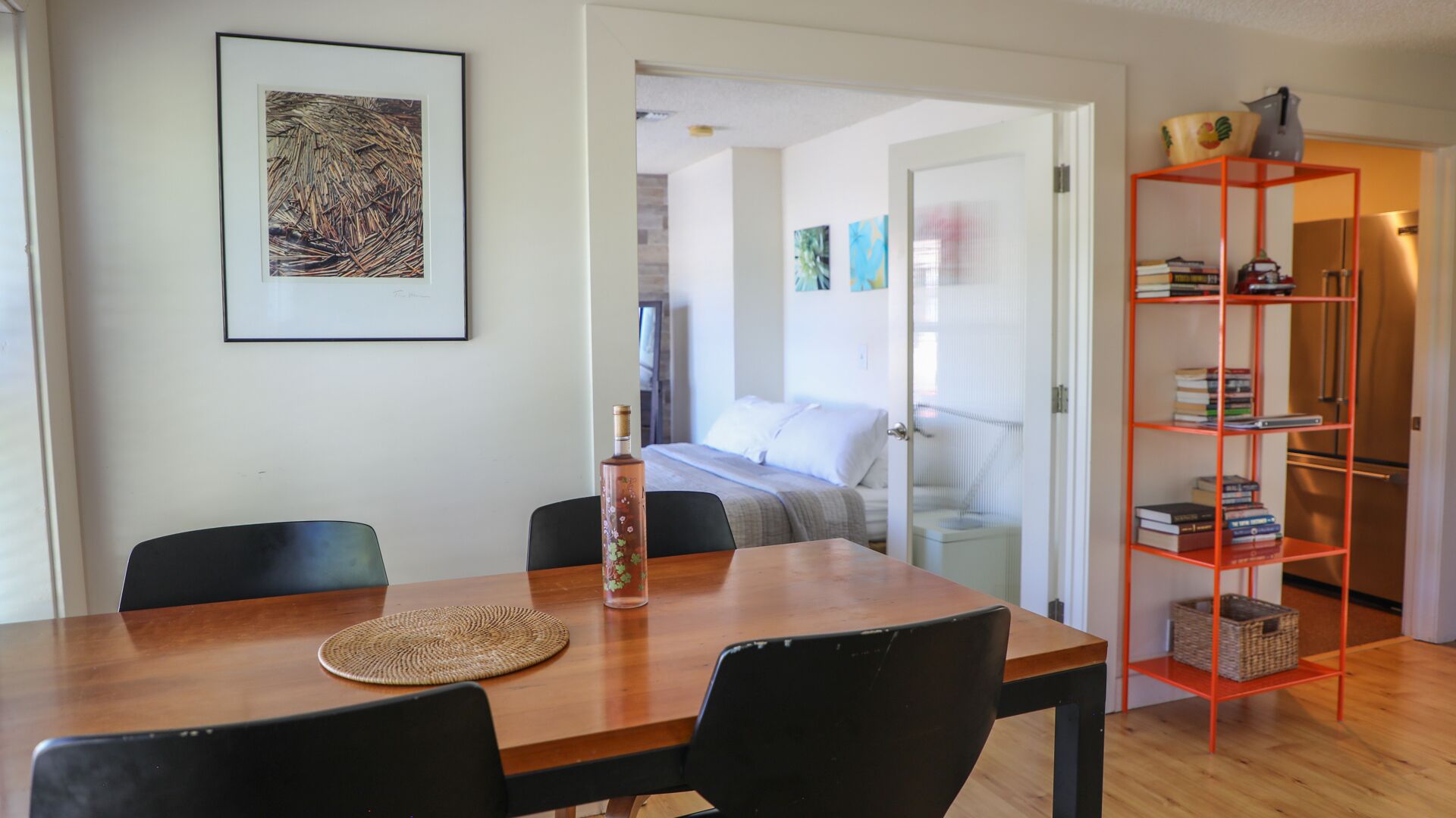 A dining area with a wooden table, chairs, a woven placemat, and a bottle. A framed abstract artwork is on the wall.