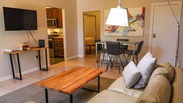 A living room with a sofa, a wooden coffee table, a TV, a kitchen area with cabinets and appliances, and a dining area with chairs and a table.