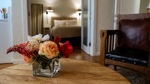 A room with a bed, a wooden chair, and a glass vase with flowers on a wooden table. - Suites On Main (Kelseyville)
