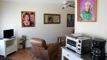A living room with a television, a brown leather chair, a white cabinet, a microwave, a coffee maker, and three framed artworks on the wall.
