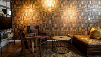 A room with a bookshelf wall, a leather sofa, a wooden chair, a small round table, and a television.