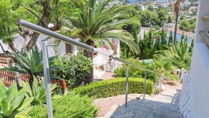 Una escalera con barandillas metálicas, rodeada de abundante vegetación y vistas a una colina con edificios.