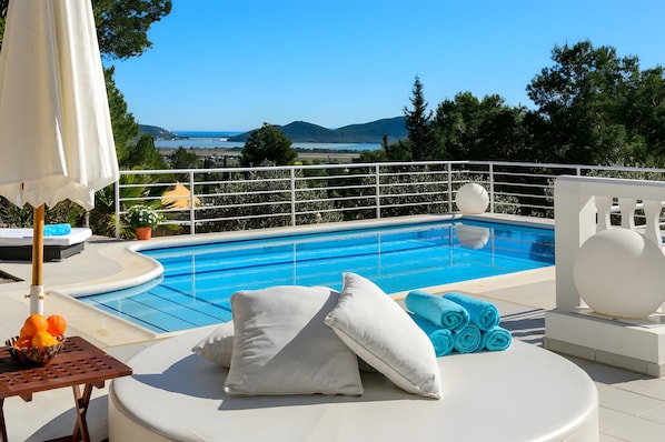 A poolside area with a view of the sea, a white lounge chair, a table with fruits, and a white umbrella.
