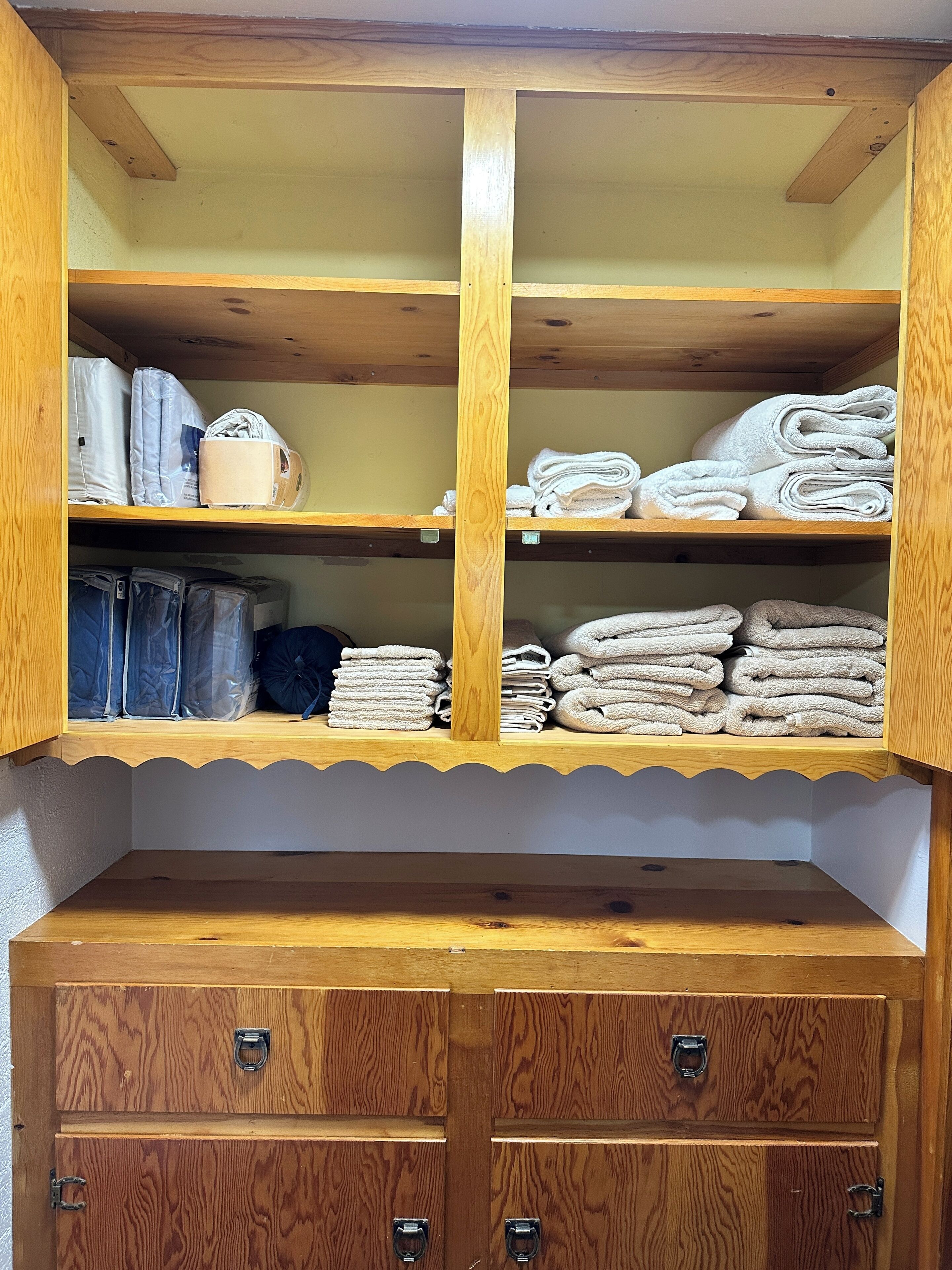 A wooden storage unit with multiple shelves and drawers, containing folded towels and plastic-wrapped items.