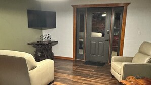 Ein Wohnzimmer mit einer Couch, einem Sessel und einem grünen Sessel. Ein Fernseher ist an der Wand montiert und eine Tür mit Glasfenster ist vorhanden.