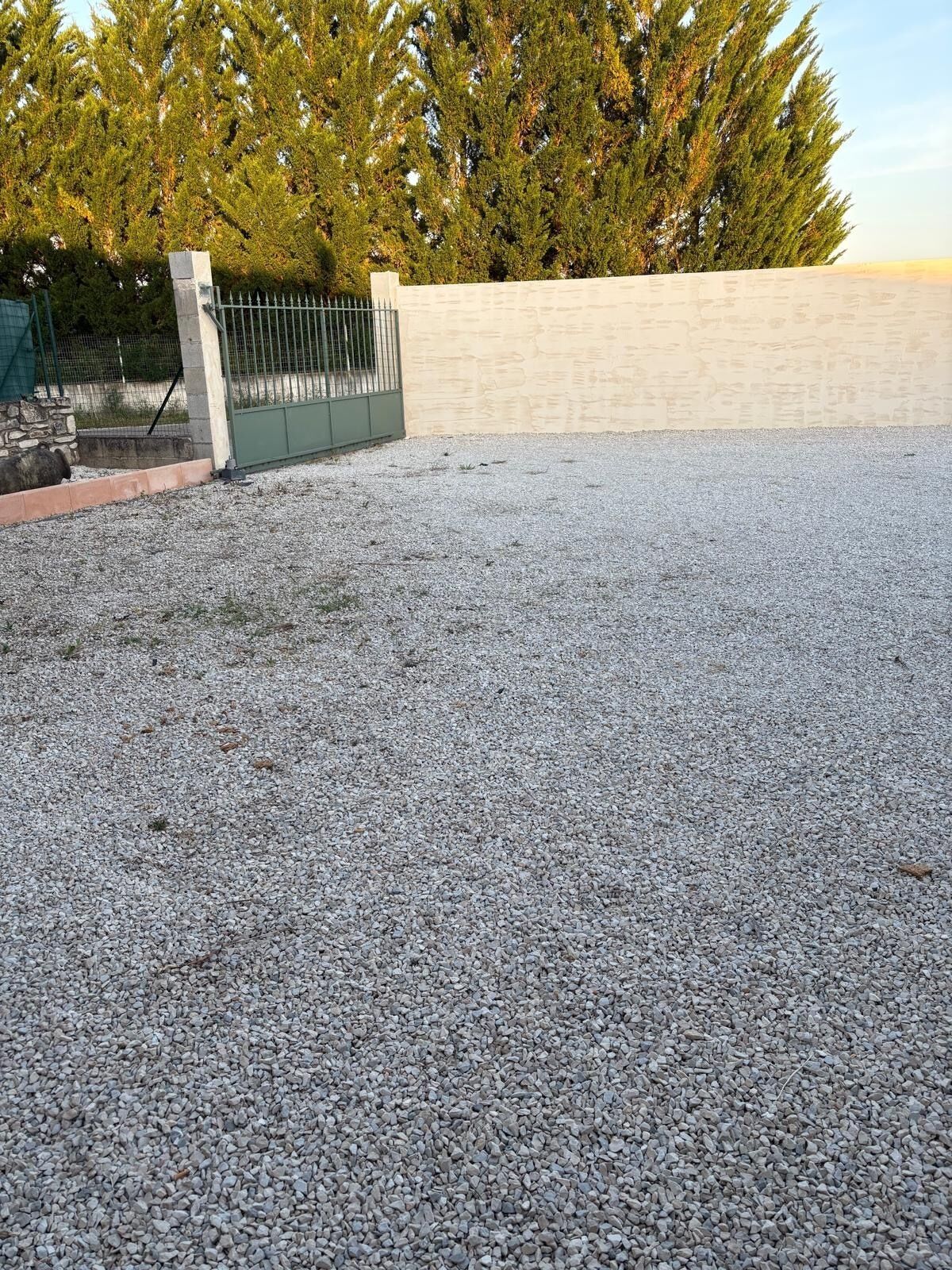 A gravel driveway leading to a gated entrance with a stone wall and trees in the background.