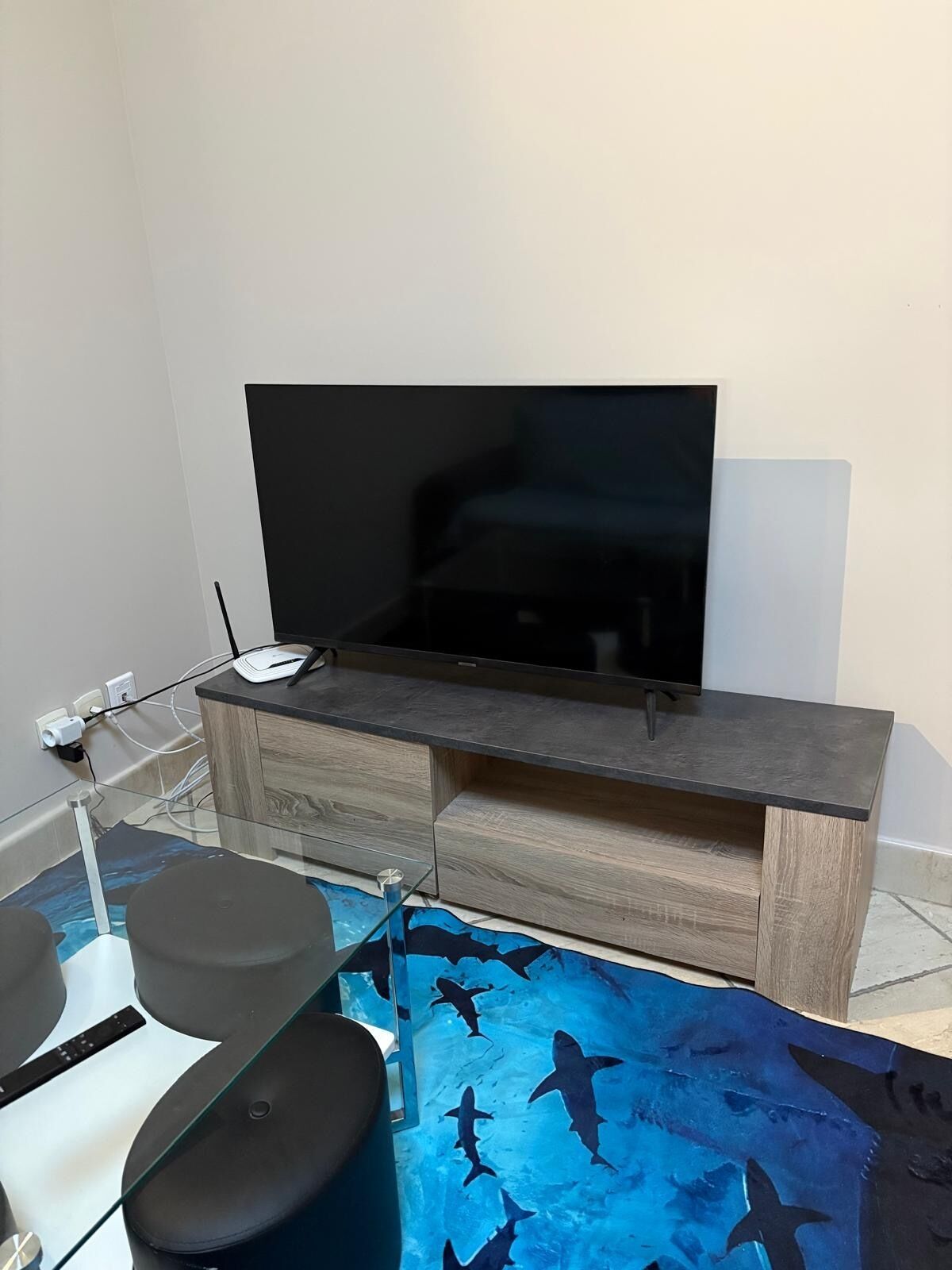 A modern living room with a flat-screen TV, wooden cabinet, glass coffee table, and a blue shark-patterned rug.