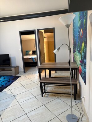 A modern living room with a wooden bench, a standing lamp, a television, and a colorful painting on the wall. - Beautiful apartment with pool access (La Rouvière)