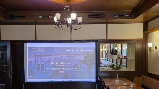 Una pantalla proyectando el sitio web de un hotel, una mesa de madera con sillas, una araña y un estante con adornos.