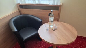 Una habitación de esquina con un sillón de cuero negro, una mesa redonda de madera con dos vasos y una botella, y una alfombra roja.