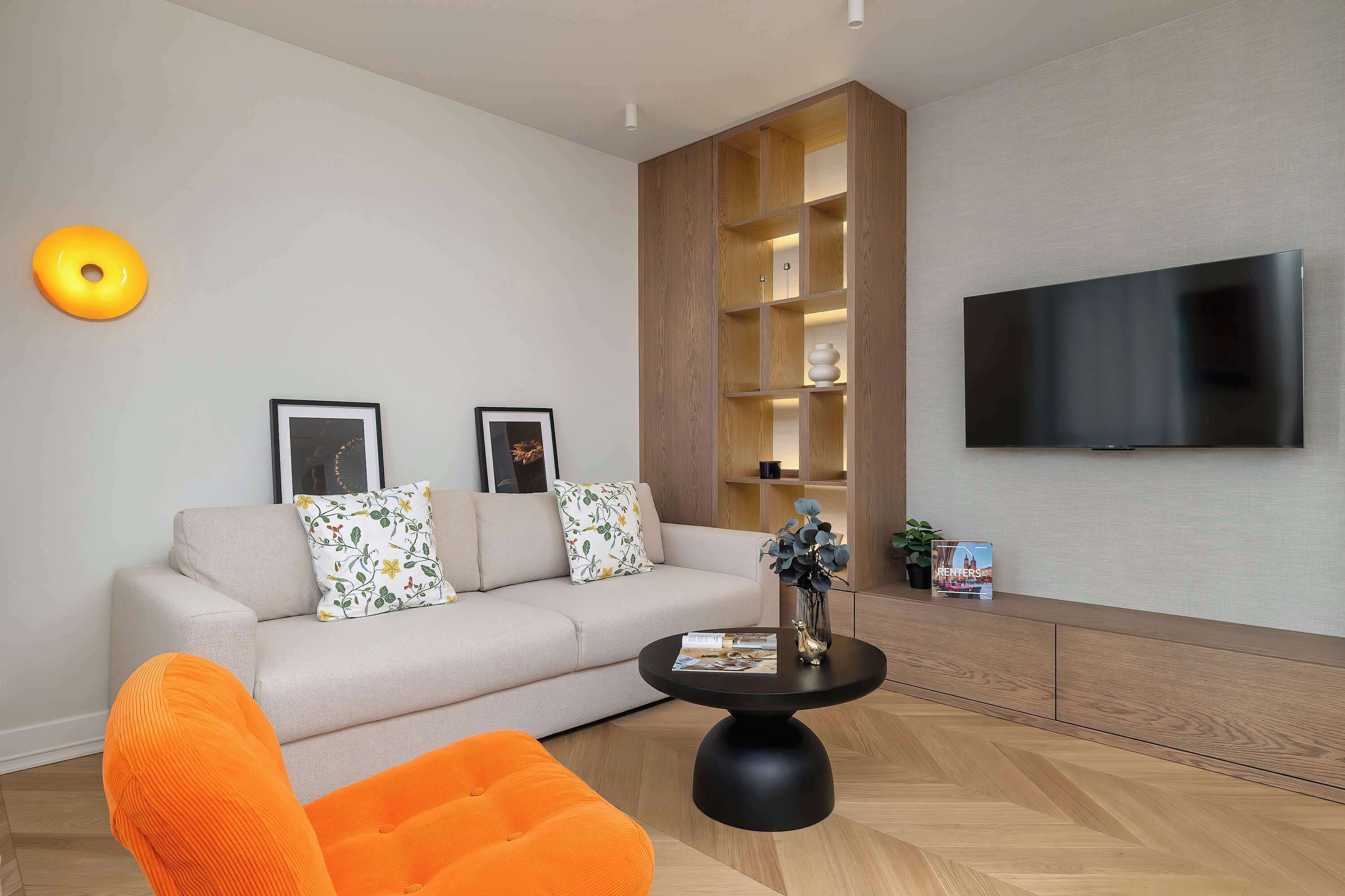 A modern living room with a sofa, a coffee table, a bright orange chair, a wooden cabinet, a flat-screen TV, and a wall-mounted light fixture.