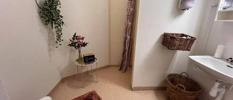 Une salle de bain avec une douche, un lavabo, une petite table avec un vase, un panier en osier et un tapis moelleux.