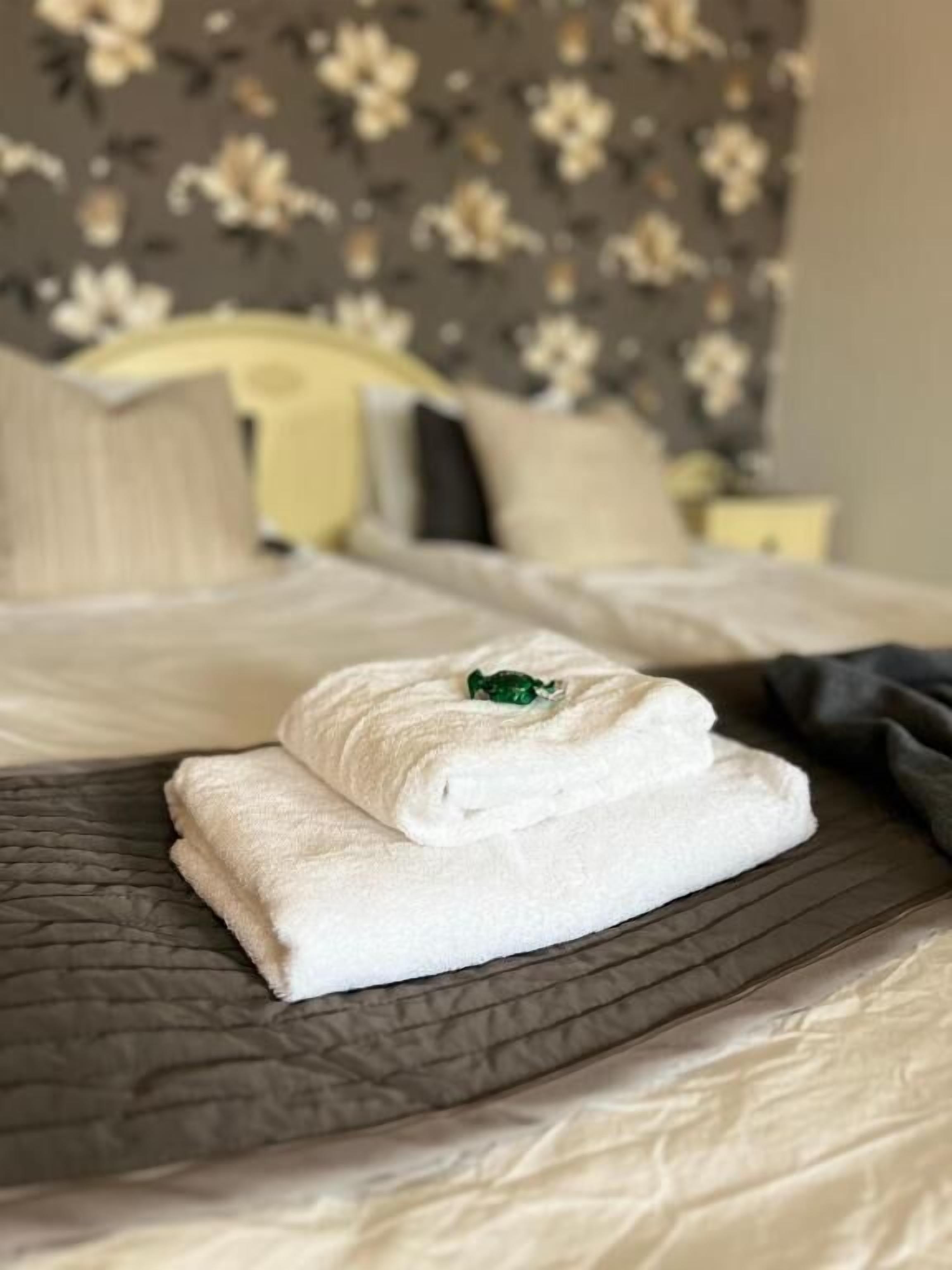 A neatly made bed with a folded white towel and a green object on top, a floral-patterned headboard, and a bedside table with a lamp.
