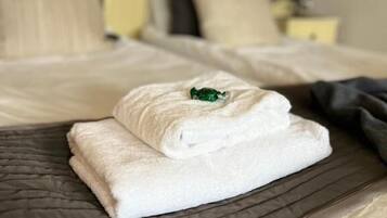 A neatly made bed with a folded white towel and a green object on top, a floral-patterned headboard, and a bedside table with a lamp.