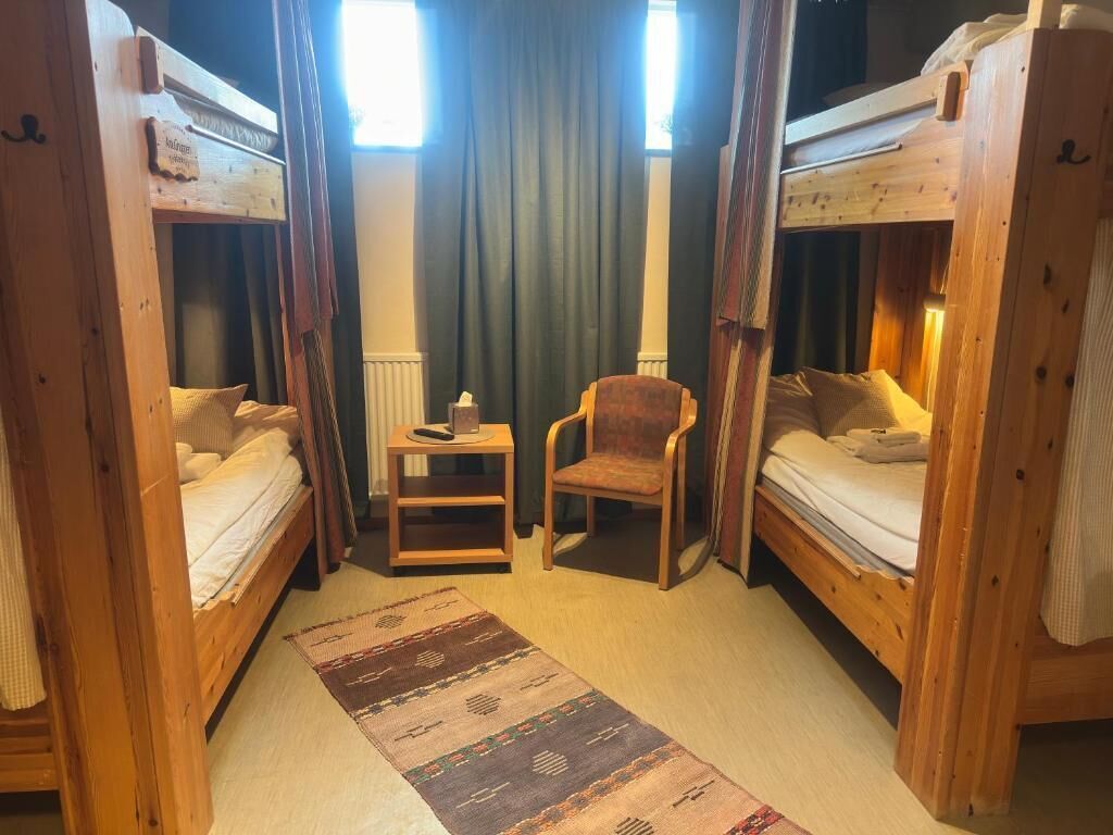 Um quarto com duas beliches de madeira, uma cadeira de madeira pequena, um tapete e uma janela com cortinas.