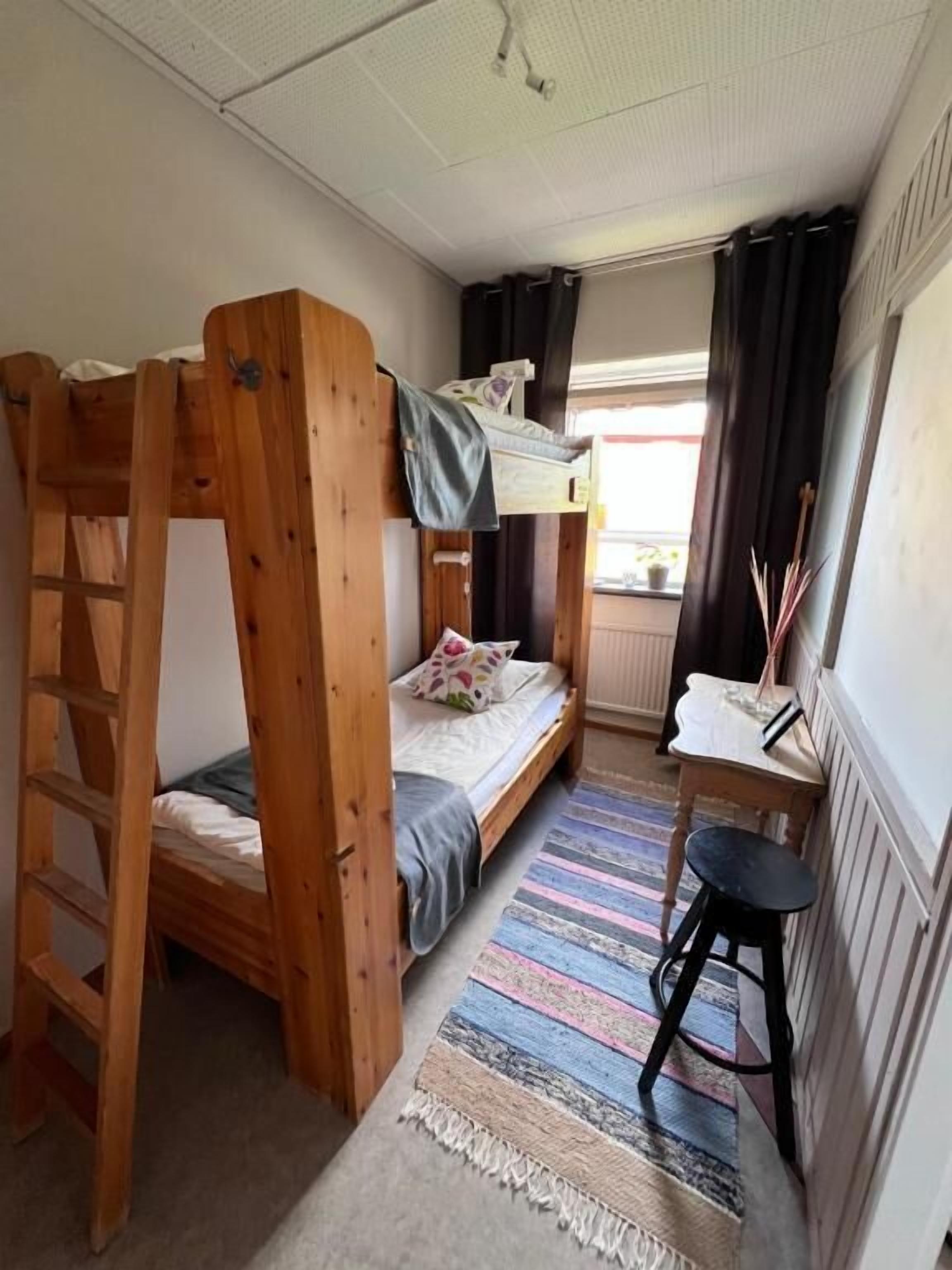 A bunk bed with a ladder, a desk with a chair, a striped rug, and a window with curtains.