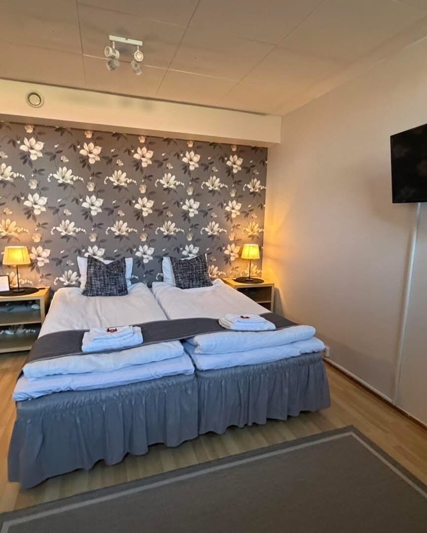 A double bed with a grey bed skirt, two bedside tables with lamps, a floral-patterned wall, and a flat-screen TV mounted on the wall.