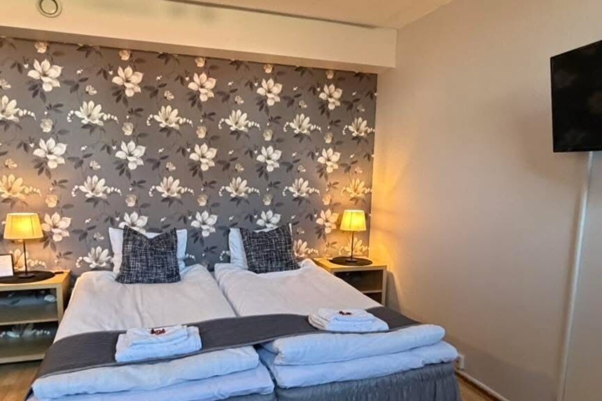 A double bed with a grey bed skirt, two bedside tables with lamps, a floral-patterned wall, and a flat-screen TV mounted on the wall.