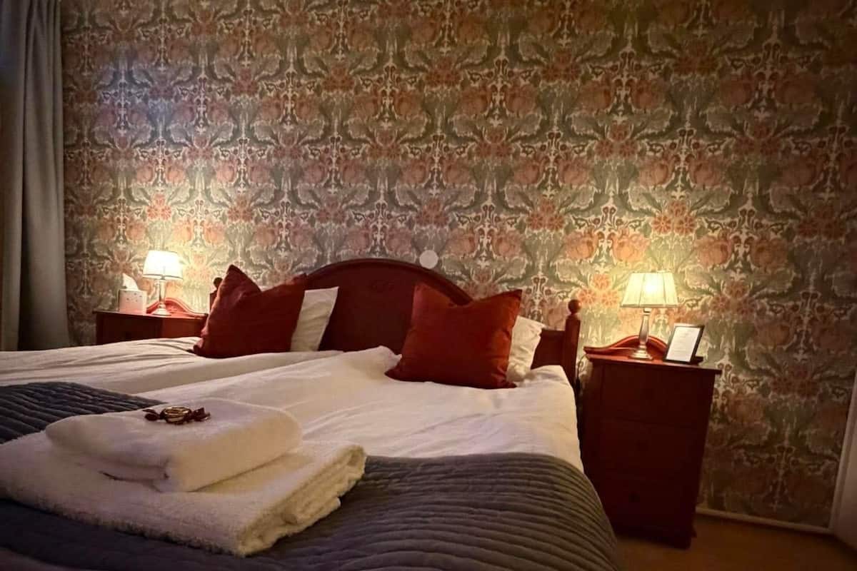 A hotel room with a large bed, two bedside tables with lamps, a patterned wallpaper, and a window with curtains.