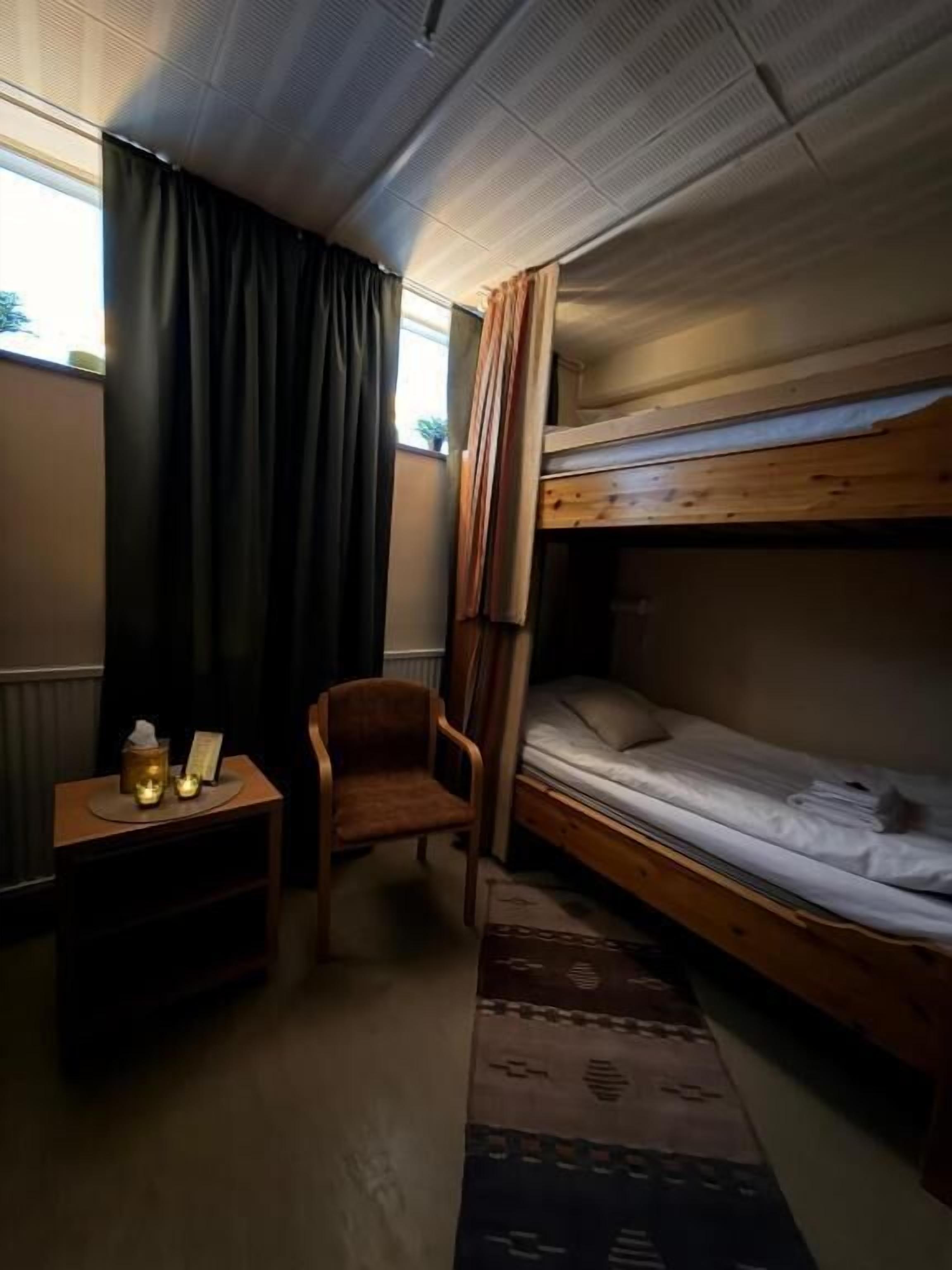 A compact room with a bunk bed, a small wooden nightstand with a lamp, a chair, and a patterned rug.