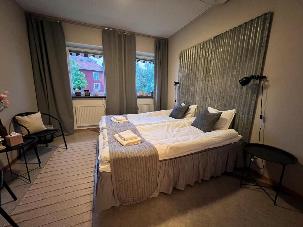 Quarto de hotel com uma cama grande, uma mesinha, uma cadeira, janela com cortinas e vista para um edifício.