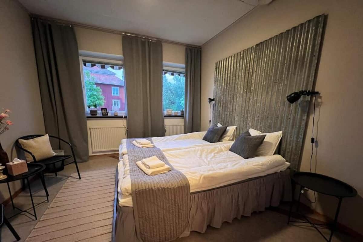 A hotel room with a large bed, a small table, a chair, a window with curtains, and a view of a building outside.