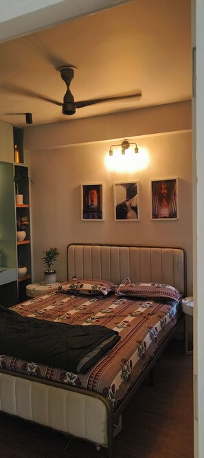 Quarto com cama, ventilador de teto, luminária na parede e prateleira com plantas.