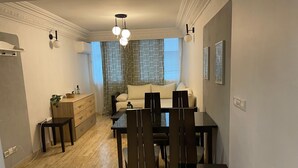 En matplats med ett bord och stolar, en soffa, ett fönster med mönstrade gardiner och en väggmonterad lampa.