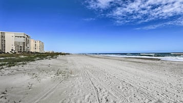 A wide sandy beach with footprints, a clear blue sky, and a building in the background.