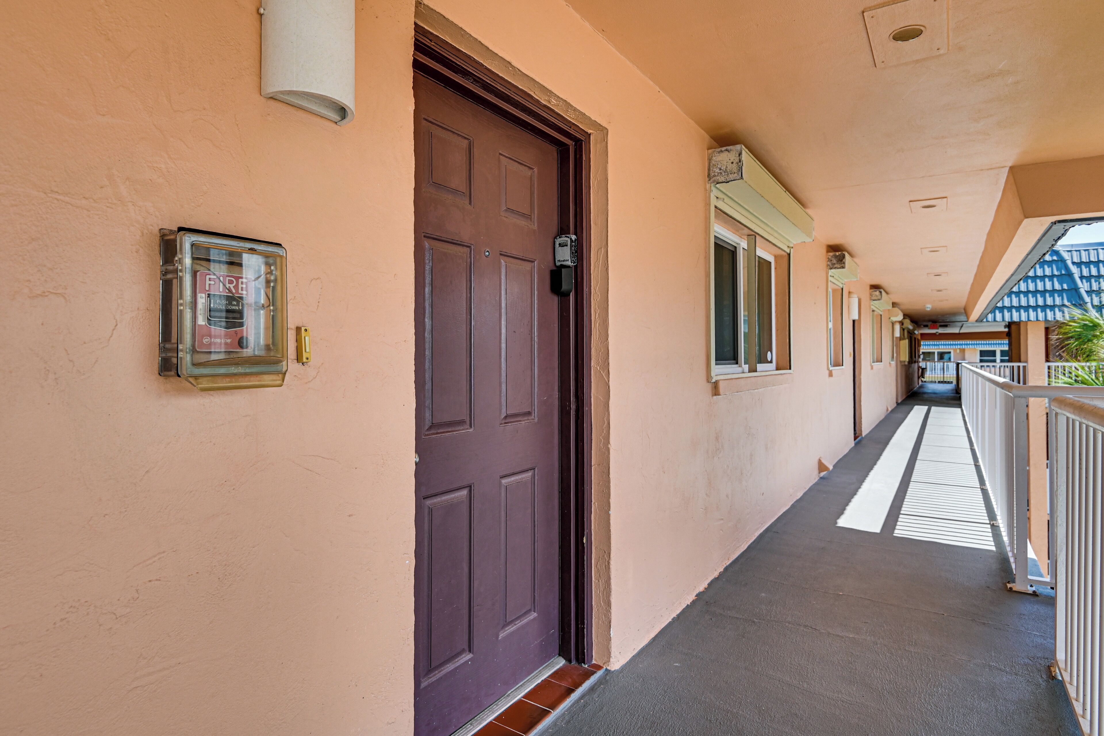 A hallway with a brown door, a fire alarm, and a window on the right.