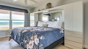 Un dormitorio con cama, ventilador de techo, ventana grande con vista a la playa y armario empotrado.