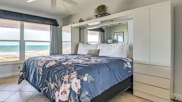 A bedroom with a bed, a ceiling fan, a large window with a view of the beach, and a built-in wardrobe.