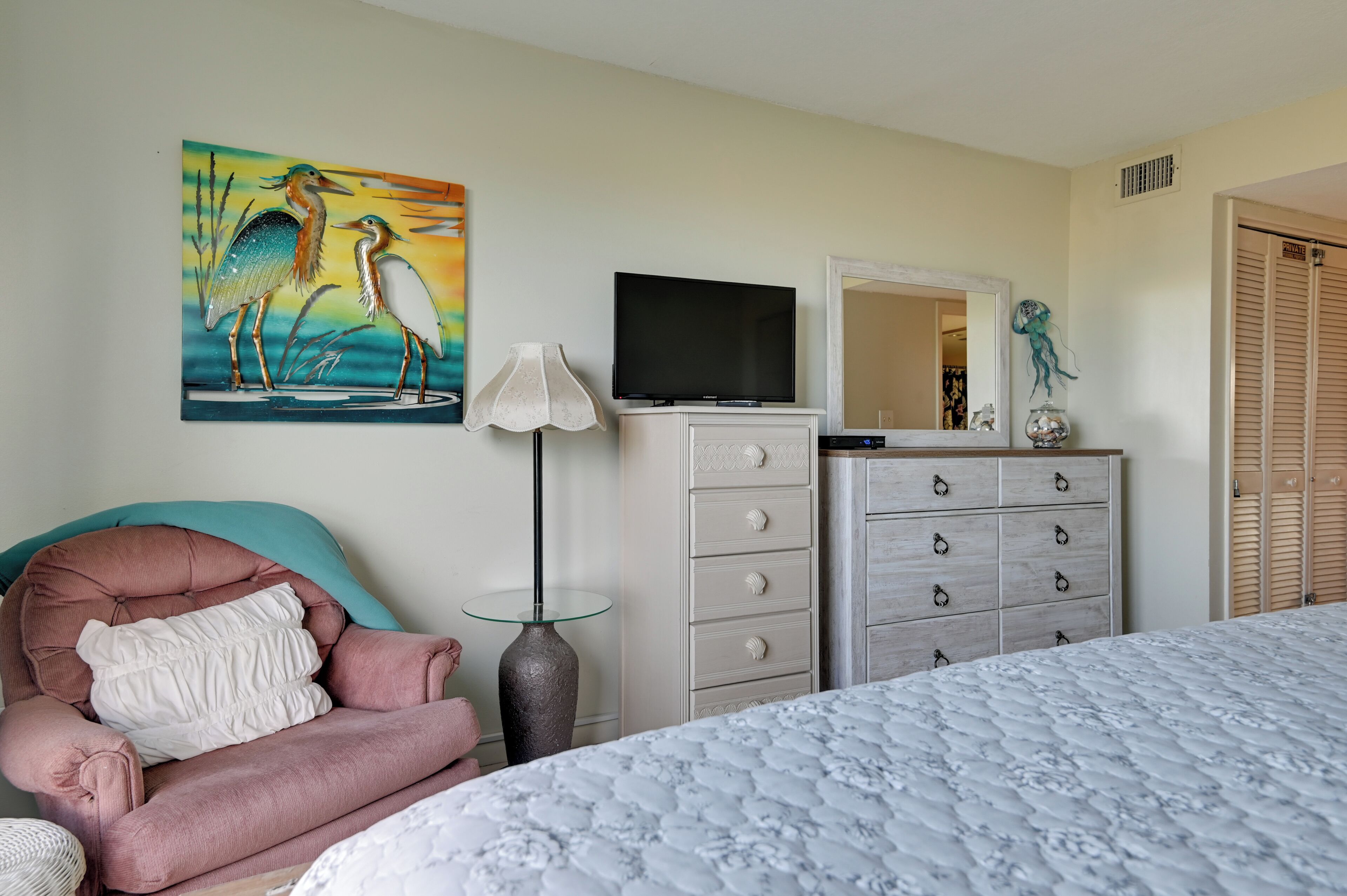 A bedroom with a bed, a TV, a dresser, a chair, a lamp, and a painting.