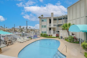 Piscina com espreguiçadeiras e guarda-sóis, edifício com varandas e céu claro.