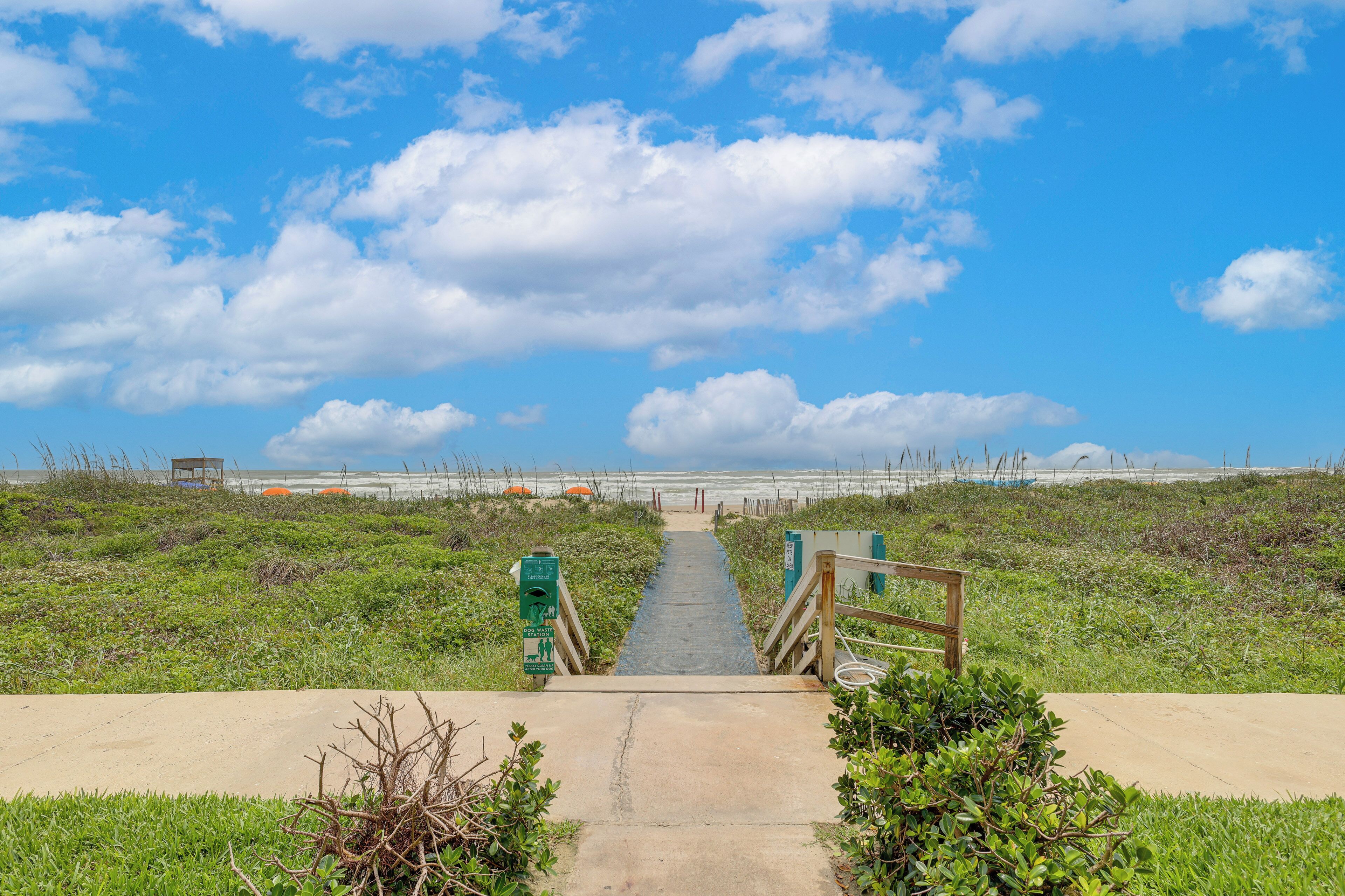 A concrete pathway leading to a beach with a wooden fence on one side and a lifeguard tower in the distance.