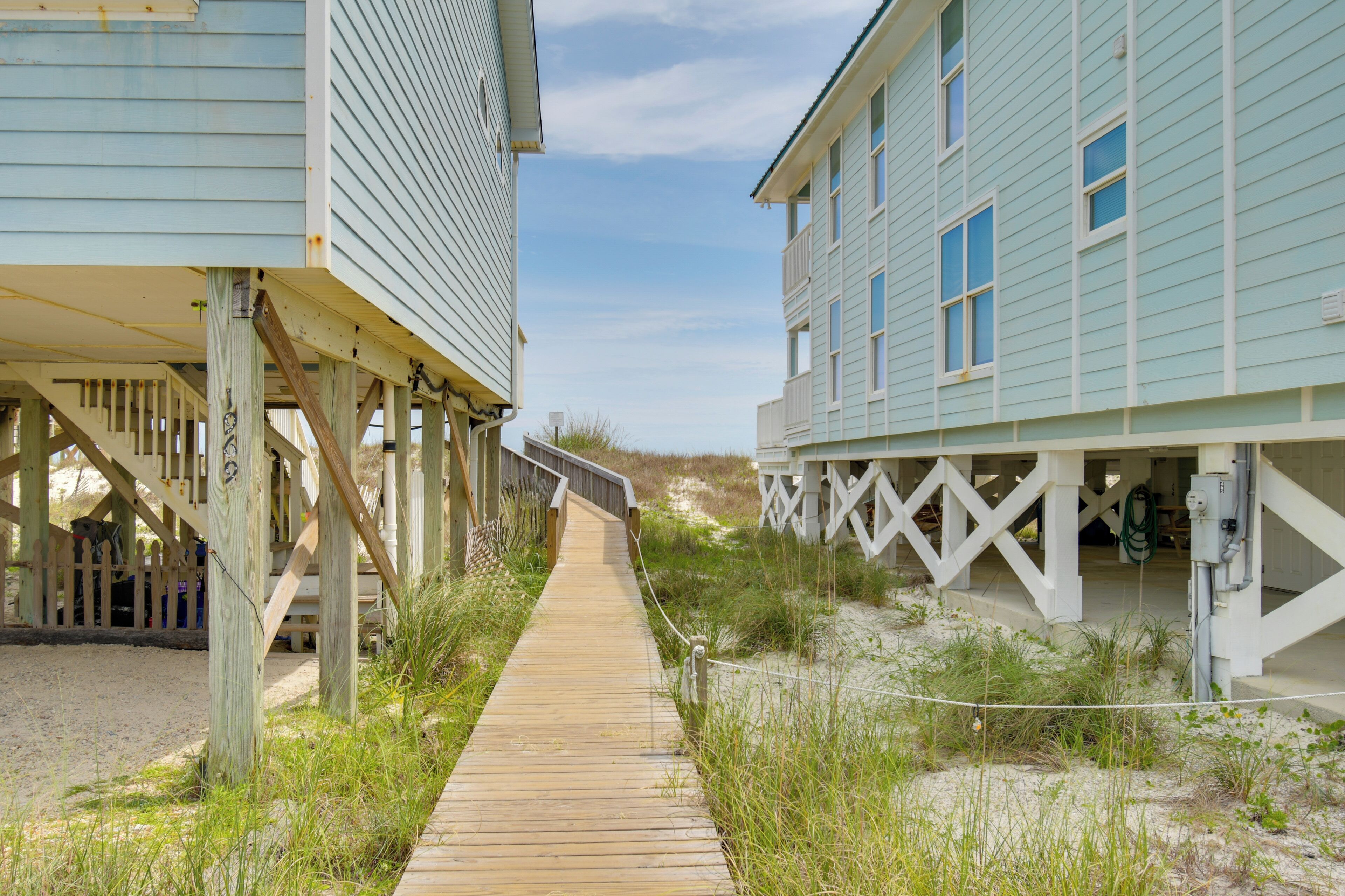 En träpromenad som leder till strandhus, med en sandstrand och sanddyner i bakgrunden.
