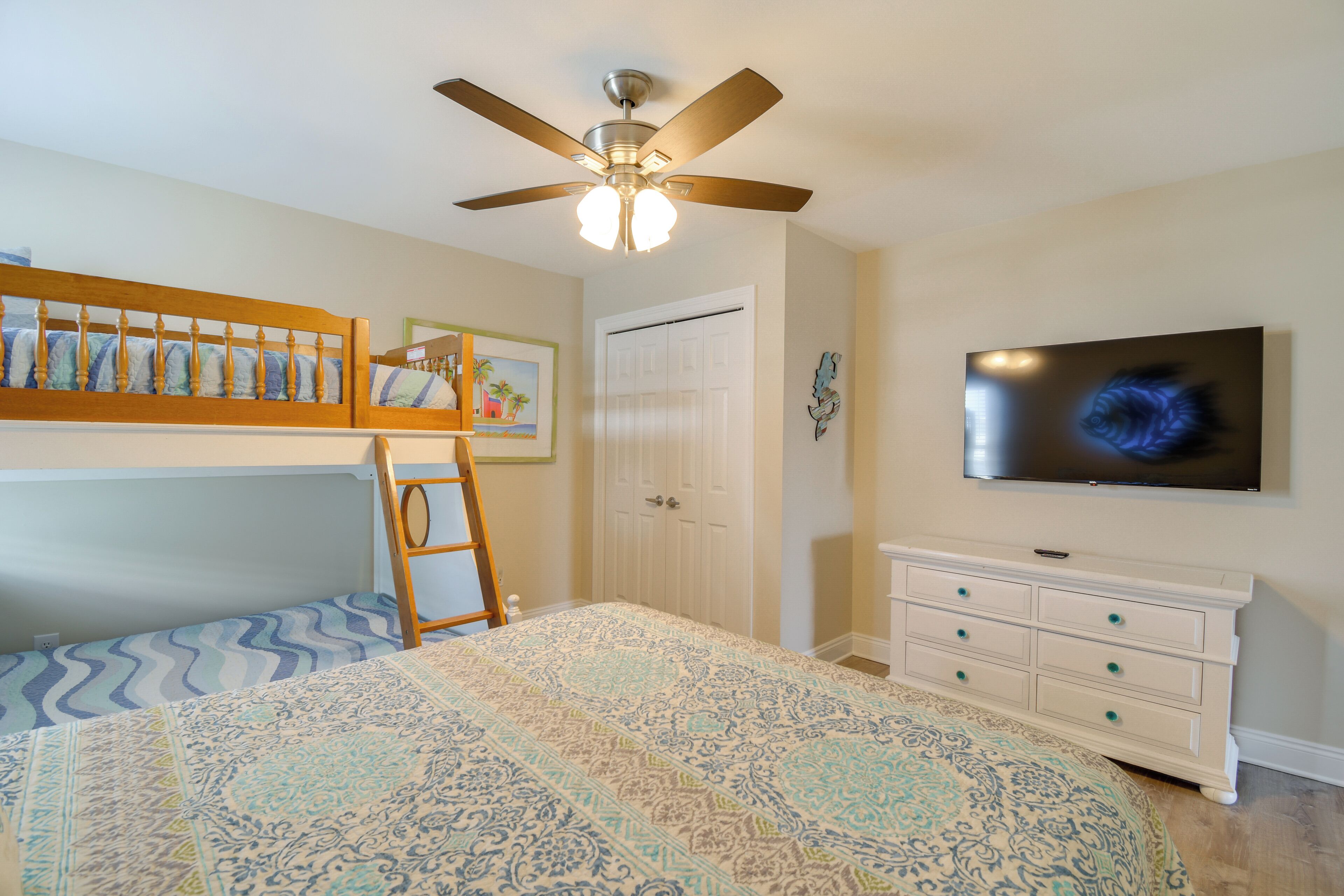 A bedroom with bunk beds, a ceiling fan, a dresser, a TV, and a door.