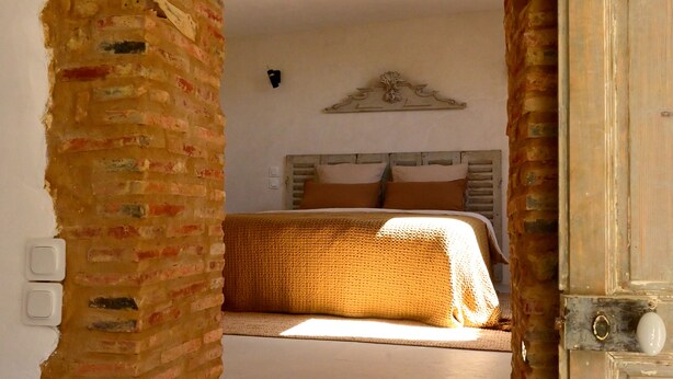 A room with a brick wall, a bed with a white headboard, and a wooden door. - Au Canari, a former bar-tabac transformed into a charming gîte in the village of Colette. (Saint-Sauveur-en-Puisaye)
