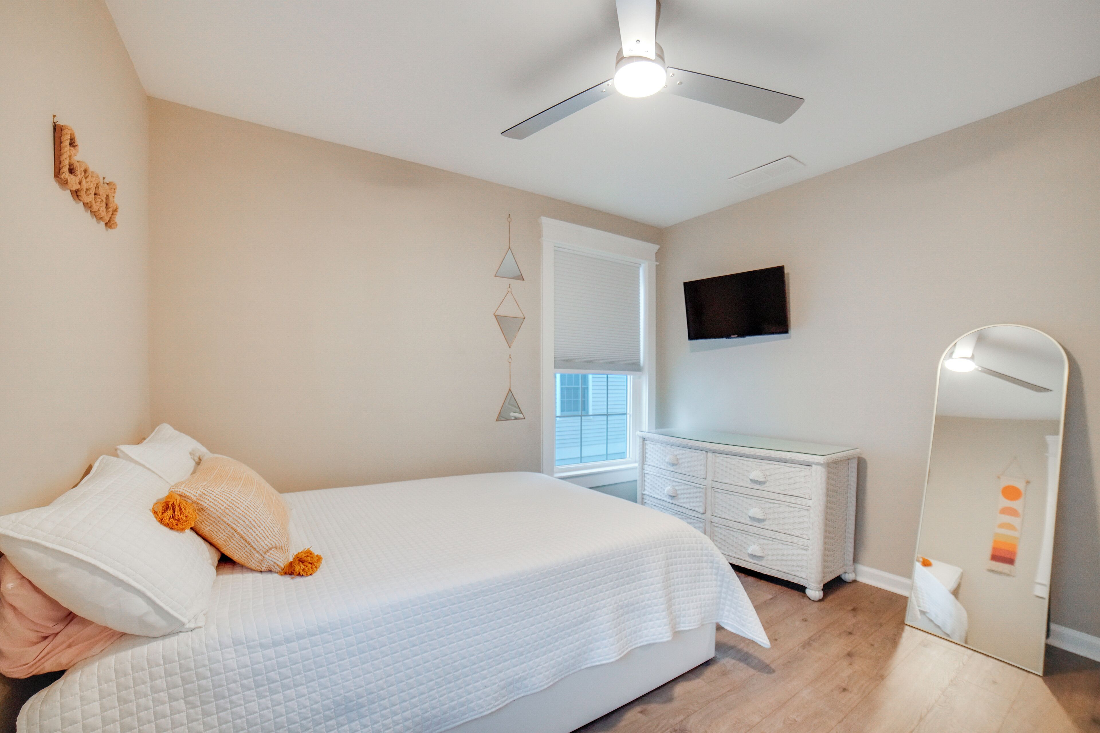 A bedroom with a bed, dresser, mirror, ceiling fan, and a TV on the wall.