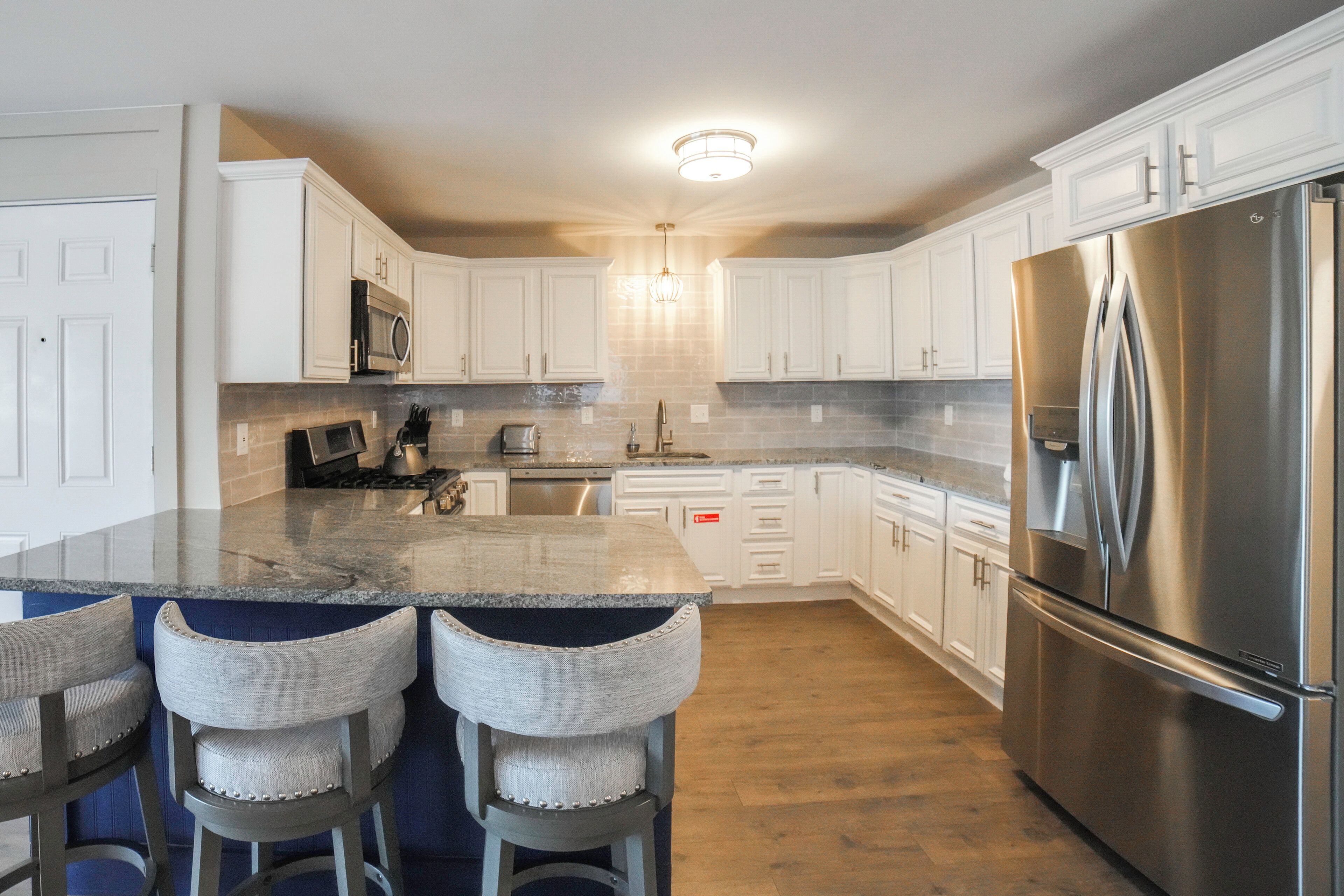 A modern kitchen with white cabinets, a stainless steel refrigerator, a granite countertop, and bar stools.